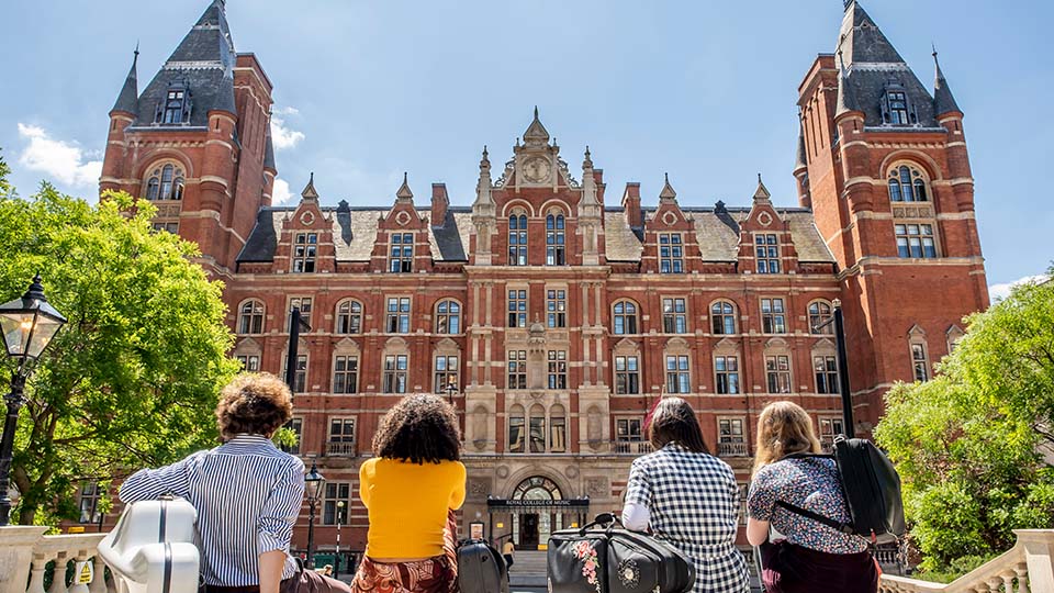 Four students sat in a line facing the RCM's Blomfield building, with blue sky and green trees